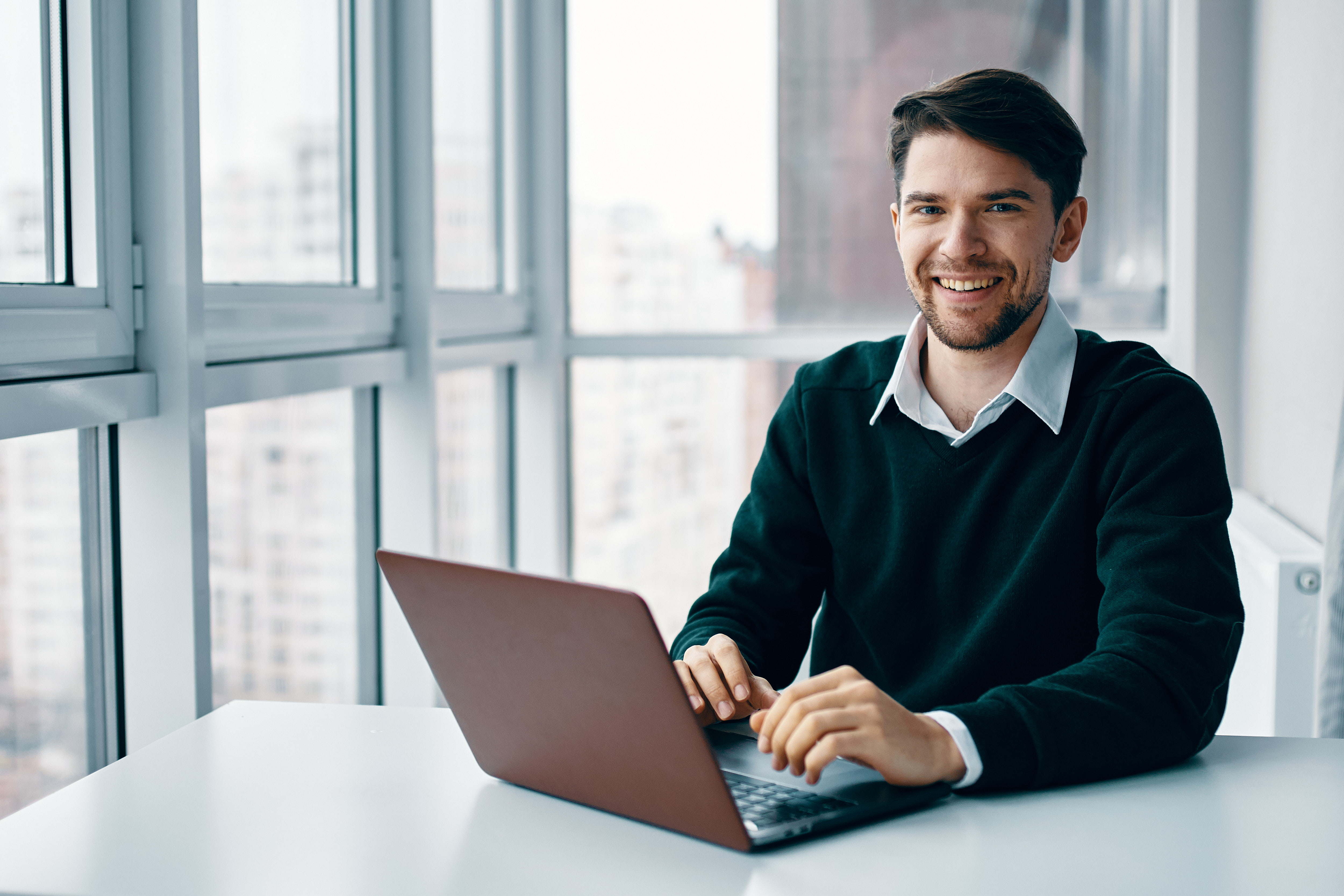 Young man with a laptop in a business suit working in the office and at home on the background of a window, interviewing online